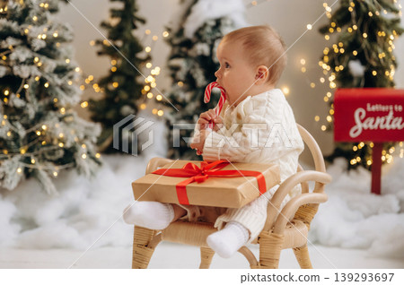 Sweet candy in hand. Little baby is sitting indoors in the Christmas decorated room Sweet candy in hand. Little baby is sitting indoors in the Christmas decorated room 139293697