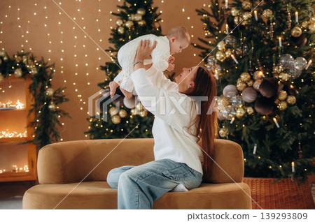 Side view, holding kid in hands. Mother is with her little baby daughter are indoors, celebrating Christmas Side view, holding kid in hands. Mother is with her little baby daughter are indoors, celebrating Christmas 139293809