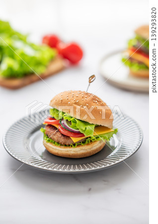 Beef burger with tomato, cheese, and lettuce on a plate on a white marble table with vegetables. Beef burger with tomato, cheese, and lettuce on a plate on a white marble table with vegetables. 139293820