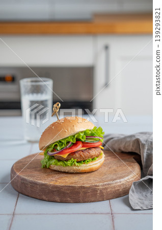 Meat burger with tomato, cheese, and lettuce on a wooden board on a table with glass of water 139293821