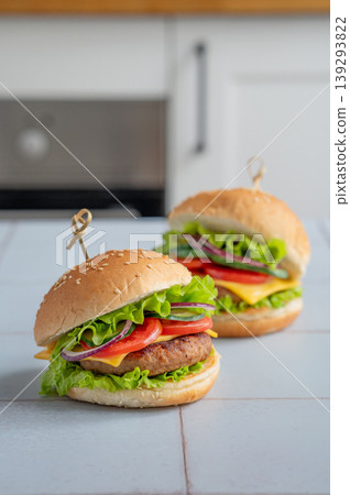 Two meat burgers with tomato, cheese, and lettuce on a table against a white kitchen Two meat burgers with tomato, cheese, and lettuce on a table against a white kitchen 139293822
