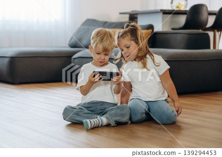 Smartphone in hands of boy. Little kids are playing together in the domestic room at daytime Smartphone in hands of boy. Little kids are playing together in the domestic room at daytime 139294453