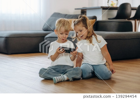 Smartphone in hands of boy. Little kids are playing together in the domestic room at daytime Smartphone in hands of boy. Little kids are playing together in the domestic room at daytime 139294454