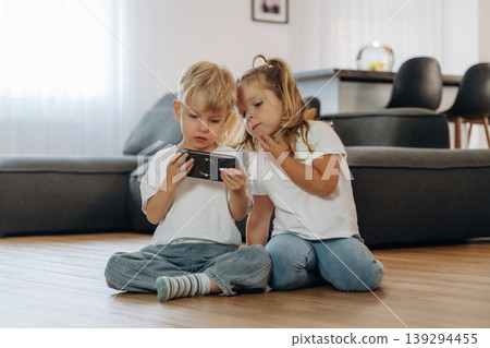 Smartphone in hands of boy. Little kids are playing together in the domestic room at daytime Smartphone in hands of boy. Little kids are playing together in the domestic room at daytime 139294455