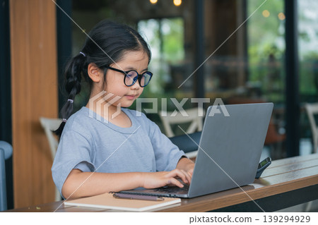 Young Girl with Glasses Engaged in Writing Notes at a Laptop in a Cozy Cafe Environment Under Soft Natural Light Young Girl with Glasses Engaged in Writing Notes at a Laptop in a Cozy Cafe Environment Under Soft Natural Light 139294929