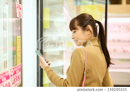 A woman shopping at a supermarket while looking at her smartphone. 139295835