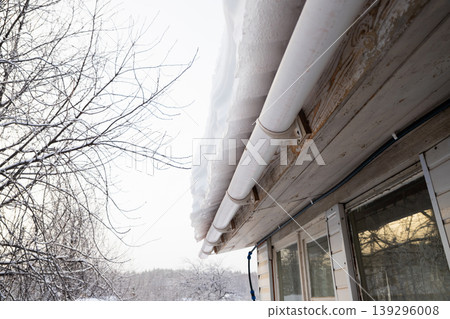 danger of snow falling from roof. Snow hangs heavily from gutter and roof edge of a house with bare winter trees in background, depicting cold weather and winter conditions danger of snow falling from roof. Snow hangs heavily from gutter and roof edge of a house with bare winter trees in background, depicting cold weather and winter conditions 139296008