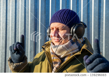 Man, who is a farmer, wearing a warm hat and scarf listening to music with headphones outside. Rural worker enjoying free time in a countryside setting. Leisure and relaxation concept. Man, who is a farmer, wearing a warm hat and scarf listening to music with headphones outside. Rural worker enjoying free time in a countryside setting. Leisure and relaxation concept. 139296083