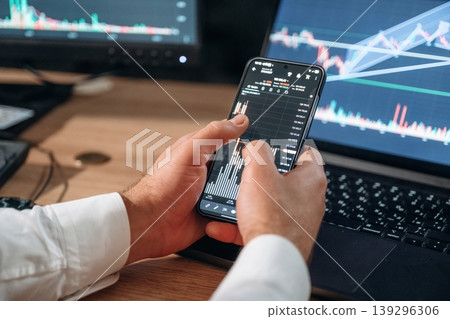 Against the laptop. Detailed close up view of man's hands that is holding smartphone with stock market crypto information Against the laptop. Detailed close up view of man's hands that is holding smartphone with stock market crypto information 139296306