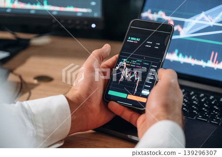 Against the laptop. Detailed close up view of man's hands that is holding smartphone with stock market crypto information Against the laptop. Detailed close up view of man's hands that is holding smartphone with stock market crypto information 139296307