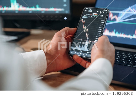 Against the laptop. Detailed close up view of man's hands that is holding smartphone with stock market crypto information Against the laptop. Detailed close up view of man's hands that is holding smartphone with stock market crypto information 139296308