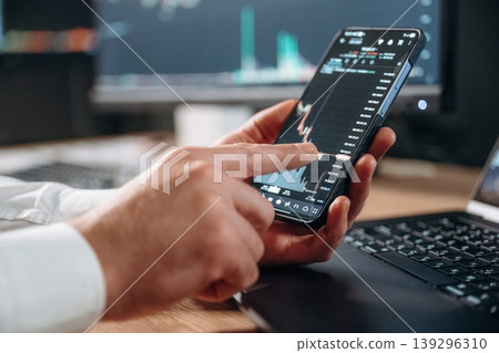 Detailed close up view of man's hands that is holding smartphone with stock market crypto information Detailed close up view of man's hands that is holding smartphone with stock market crypto information 139296310