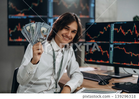 Rich, profit, holding currency in hand. Young man is sitting in the office with multiple displays of stock market data Rich, profit, holding currency in hand. Young man is sitting in the office with multiple displays of stock market data 139296318