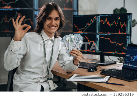 Rich, profit, holding currency in hand. Young man is sitting in the office with multiple displays of stock market data Rich, profit, holding currency in hand. Young man is sitting in the office with multiple displays of stock market data 139296319