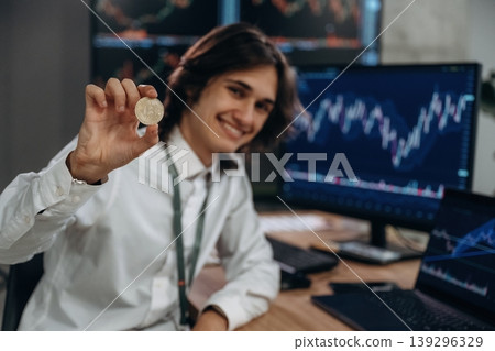 Investment concept, holding coin of crypto currency. Young man is sitting in the office with multiple displays of stock market data Investment concept, holding coin of crypto currency. Young man is sitting in the office with multiple displays of stock market data 139296329