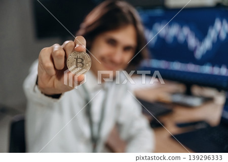 Golden colored crypto coin. Young man is sitting in the office with multiple displays of stock market data Golden colored crypto coin. Young man is sitting in the office with multiple displays of stock market data 139296333