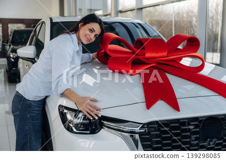 Red gift ribbon on a car. Woman posing with a car in an auto showroom, modern dealership environment 139298085