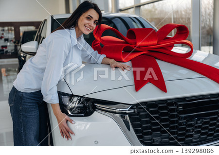 Red gift ribbon on a car. Woman posing with a car in an auto showroom, modern dealership environment 139298086