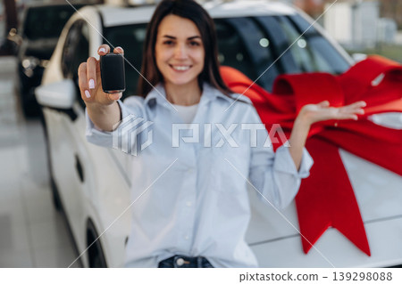 Red gift ribbon on a car. Woman posing with a car in an auto showroom, modern dealership environment 139298088
