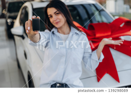Red gift ribbon on a car. Woman posing with a car in an auto showroom, modern dealership environment 139298089