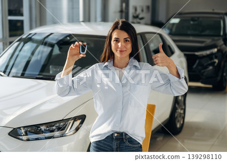 Brand new vehicle. Woman posing with a car in an auto showroom, modern dealership environment Brand new vehicle. Woman posing with a car in an auto showroom, modern dealership environment 139298110