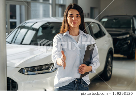 Giving hand to shake. Woman posing with a car in an auto showroom, modern dealership environment Giving hand to shake. Woman posing with a car in an auto showroom, modern dealership environment 139298120