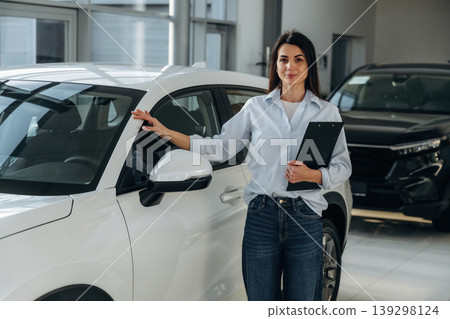 Brand new vehicle. Woman posing with a car in an auto showroom, modern dealership environment Brand new vehicle. Woman posing with a car in an auto showroom, modern dealership environment 139298124