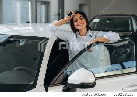 Smiling, happy new owner. Woman posing with a car in an auto showroom, modern dealership environment 139298133