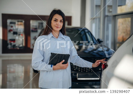 Notepad is in hands. Woman posing with a car in an auto showroom, modern dealership environment Notepad is in hands. Woman posing with a car in an auto showroom, modern dealership environment 139298407
