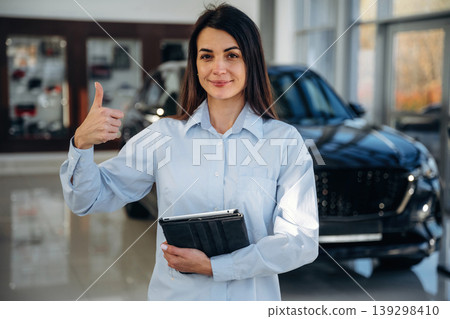 Manager is standing. Woman posing with a car in an auto showroom, modern dealership environment Manager is standing. Woman posing with a car in an auto showroom, modern dealership environment 139298410