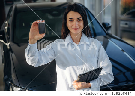 Manager is standing. Woman posing with a car in an auto showroom, modern dealership environment Manager is standing. Woman posing with a car in an auto showroom, modern dealership environment 139298414