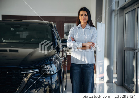 Looking at the camera. Woman posing with a car in an auto showroom, modern dealership environment 139298424