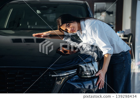 Embracing the vehicle. Woman posing with a car in an auto showroom, modern dealership environment Embracing the vehicle. Woman posing with a car in an auto showroom, modern dealership environment 139298434
