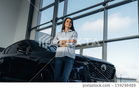 Big panoramic window is behind. Woman posing with a car in an auto showroom, modern dealership environment Big panoramic window is behind. Woman posing with a car in an auto showroom, modern dealership environment 139298489