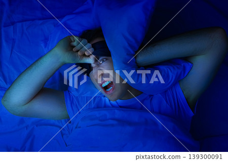 An angry man with a sleep mask covers his ears with a pillow, suffering from insomnia, noise, and sleep deprivation in a dark blue bedroom at night. An angry man with a sleep mask covers his ears with a pillow, suffering from insomnia, noise, and sleep deprivation in a dark blue bedroom at night. 139300091