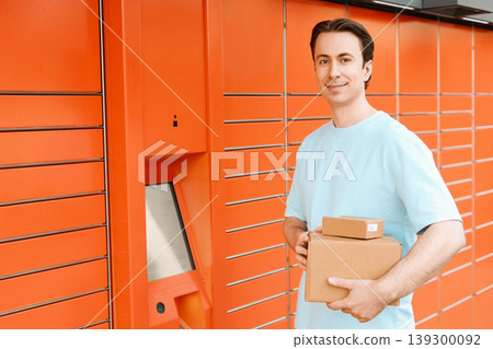 Smiling man holding cardboard parcels in front of an automated orange parcel locker, representing convenient package delivery, post service, and ecommerce shopping. Smiling man holding cardboard parcels in front of an automated orange parcel locker, representing convenient package delivery, post service, and ecommerce shopping. 139300092