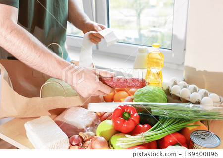 A man checks a grocery receipt while unpacking fresh food from a paper bag, including vegetables, meat, sausage, and eggs on a bright kitchen table. A man checks a grocery receipt while unpacking fresh food from a paper bag, including vegetables, meat, sausage, and eggs on a bright kitchen table. 139300096