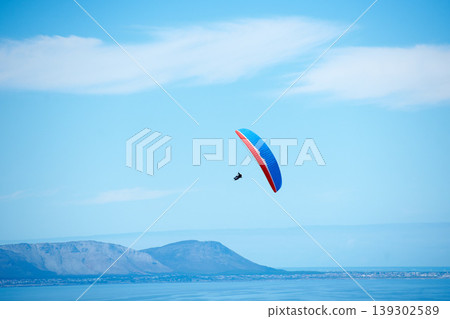 Pilot, parachute and paragliding in blue sky for flight, freedom and courage with extreme sport. Athlete, glide and fearless in outdoor fitness for adventure, health and sports by ocean with mountain Pilot, parachute and paragliding in blue sky for flight, freedom and courage with extreme sport. Athlete, glide and fearless in outdoor fitness for adventure, health and sports by ocean with mountain 139302589