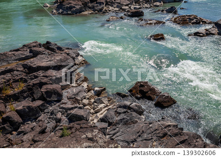 Sunlit turquoise river rapids flow around dark basalt rocks Sunlit turquoise river rapids flow around dark basalt rocks 139302606