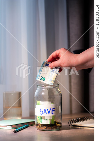 Close up of cash being added to transparent money jar. Represents traditional saving methods and conscious money management for future security. Vertical image Close up of cash being added to transparent money jar. Represents traditional saving methods and conscious money management for future security. Vertical image 139303314