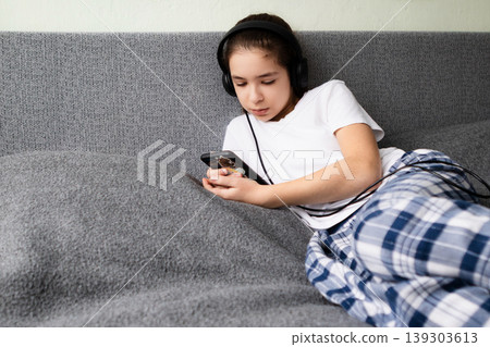 Teen girl lying on couch with headphones listening to music on smartphone in pajamas 139303613