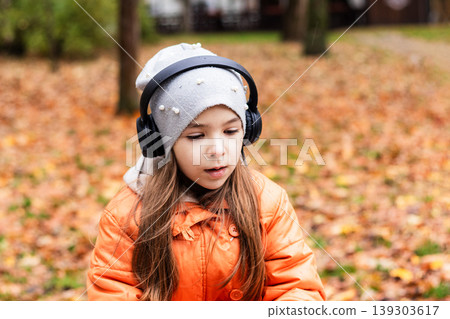 Young girl with headphones in orange coat listening outdoors among autumn leaves Young girl with headphones in orange coat listening outdoors among autumn leaves 139303617