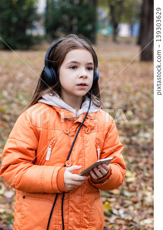 Young girl listening to music with headphones and phone in autumn park wearing orange coat Young girl listening to music with headphones and phone in autumn park wearing orange coat 139303629
