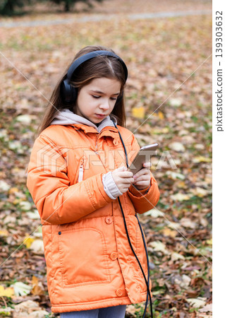 Young girl wearing headphones and orange jacket using a smartphone in an autumn park Young girl wearing headphones and orange jacket using a smartphone in an autumn park 139303632