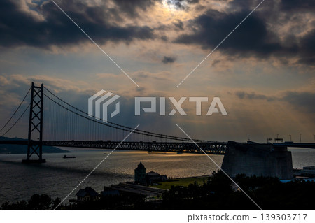 [Spectacular Sunset View] The silhouette of the Akashi Kaikyo Bridge over the beach in Maiko, Kobe, with angel's ladders of sunlight streaming down. 139303717