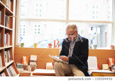 Senior man, library and reading book for literature, knowledge or story at bookstore. Mature male person, author or bookworm sitting by bookshelf for novel, learning or wisdom in shopping or leisure 139304450