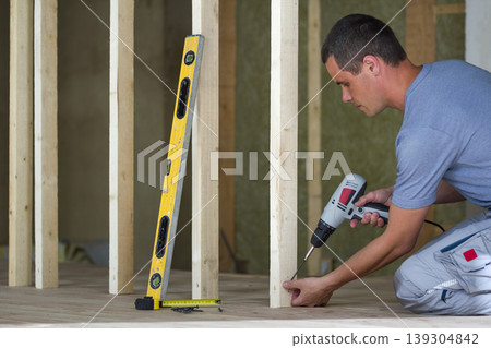 Interior of attic insulated room with oak floor under reconstruction. Young professional worker uses level and screwdriver installing wooden frame for future walls. Renovation and improvement concept. Interior of attic insulated room with oak floor under reconstruction. Young professional worker uses level and screwdriver installing wooden frame for future walls. Renovation and improvement concept. 139304842