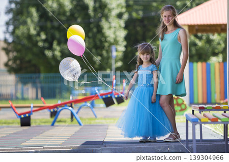 Full-length portrait of young blond smiling happily mother and small pretty daughter girl in long blue evening dress with bright colorful balloons on kindergarten playground. Care and love concept. Full-length portrait of young blond smiling happily mother and small pretty daughter girl in long blue evening dress with bright colorful balloons on kindergarten playground. Care and love concept. 139304966