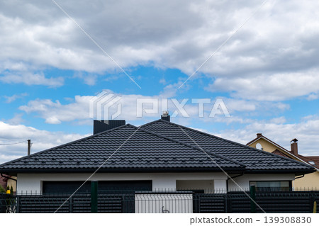 The house features a black metal tile roof, located in a residential area with a blue sky and clouds in the background The house features a black metal tile roof, located in a residential area with a blue sky and clouds in the background 139308830