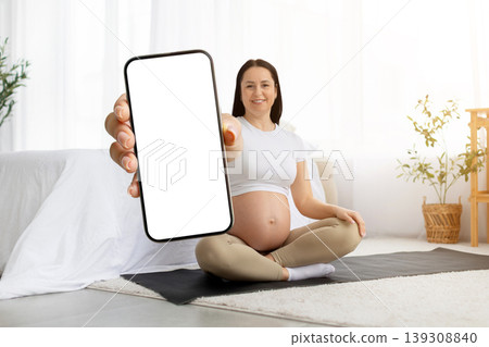 Pregnant young woman sitting on yoga mat holding smartphone with blank screen Pregnant young woman sitting on yoga mat holding smartphone with blank screen 139308840
